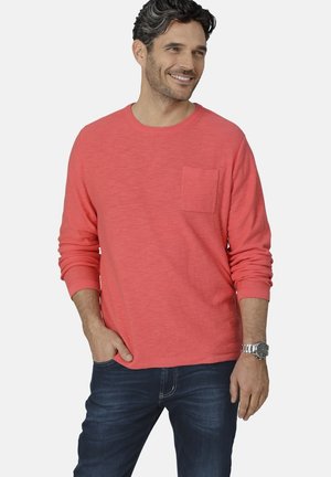 Korallenfarbener Langarm-T-Shirt mit rundem Ausschnitt, einer Brusttasche und strukturiertem Stoff. Kombiniert mit dunklen Jeans und einer silbernen Uhr.