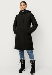 Zwarte lange jas met een capuchon, met een rechte snit, ritszakken en een gladde textuur. Gecombineerd met zwarte laarzen en blauwe jeans.