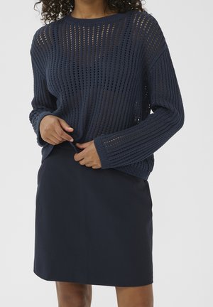 Femme portant un pull bleu marine en maille ajourée sur une jupe bleu marine foncé, ajustant l’ourlet avant du pull avec ses deux mains.