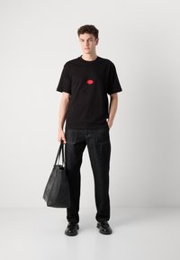 Black t-shirt with red oval logo, dark denim pants, and a structured black tote bag. Model stands on a light background, wearing black shoes.