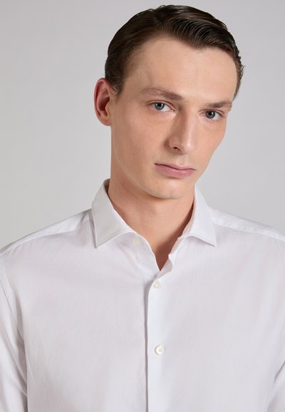 Camisa blanca de botones con cuello clásico, textura suave y corte ajustado. Presenta una tapeta frontal con botones y un diseño limpio y minimalista.