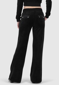 Schwarze ausgestellte Hosen aus weichem Material, mit Rückentaschen mit Druckknopfverschlüssen und einer glatten Textur. Der Bund ist hinten sichtbar.