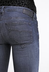 Jeans de mezclilla oscura con una textura suave, diseño ajustado y dos bolsillos traseros con costuras en ángulo y un parche de cuero en la cintura.
