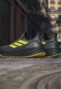 Scarpe sportive nere con accenti gialli e tre strisce, dotate di una parte superiore texture, una suola intermedia spessa e una suola con motivo.