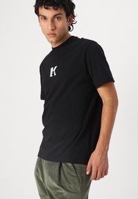 Zwarte katoenen t-shirt met een ronde hals en korte mouwen, voorzien van een wit "K"-logo op de borst. De stof heeft een soepele textuur.