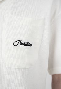 Gros plan d'une poche de chemise blanche texturée à manches courtes avec le mot « Fascinated » brodé en noir sur la partie gauche de la poitrine.