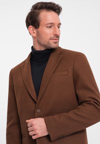 Brun ullkappa med snibbkrage, enkel knäppning och bröstficka, lager på lager över en svart ribbad turtleneck.