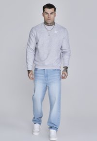 Světle šedý crewneck svetr s žebrovanými manžetami, nošený s světle modrými širokými džíny a bílými teniskami. Tetování viditelné na rukou.