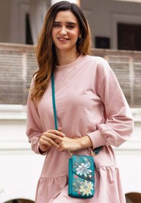 Teal leren schoudertas met bloemontwerpdetailles, gouden hardware en een verstelbare strap, gecombineerd met een roze sweatshirtjurk.