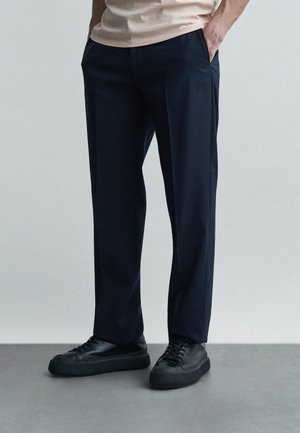 Man in zwarte, rechtgesneden broek en zwarte sneakers, staand met handen in de zakken tegen een effen achtergrond.