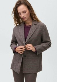 Blazer à carreaux marron avec un col à revers, en tissu texturé. Porté sur un haut bordeaux, avec une fermeture à un bouton.