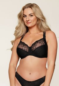 Soutien-gorge en dentelle noire avec des panneaux transparents, des motifs floraux, des bretelles réglables et un nœud décoratif au centre avant.