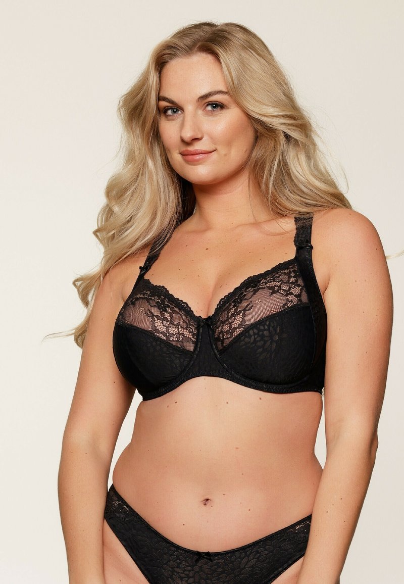 Soutien-gorge en dentelle noire avec des panneaux transparents, des motifs floraux, des bretelles réglables et un nœud décoratif au centre avant.