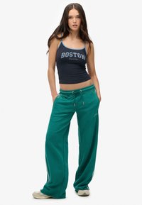 Jonge vrouw met lang bruin haar, gekleed in een marineblauwe tanktop met de tekst "Boston" en groene wide-leg sweatpants met sneakers.