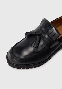 Mocassin en cuir noir avec des pampilles, un détail en frange et une épaisse semelle en caoutchouc sur un fond clair.