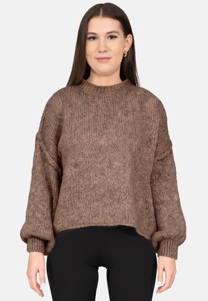 Young woman with long brown hair wearing a loose brown knit sweater and black pants, standing against a plain light background.