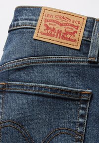 Jean en denim de couleur bleu foncé, avec des coutures distinctives sur les poches arrière, une étiquette en cuir marron clair avec un logo et un texte imprimés en rouge, et des accents de fil orange.