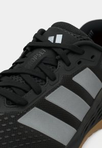 Μαύρο αθλητικό παπούτσι με πλέξη στο επάνω μέρος, με γκρι τρεις ρίγες, υφή σε διάφορα σημεία και εμφανή κορδόνια. Ονομασία μοντέλου ορατή.