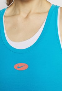 Camiseta sin mangas color turquesa hecha de algodón con un escote redondo. Presenta un logo de Nike en color rosa en el centro. Capas con una camiseta blanca debajo.