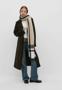Lungo cappotto marrone sopra un maglione bianco a collo alto, abbinato a jeans larghi blu e una sciarpa a motivi beige, grigia e nera.