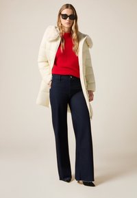 Roomwitte donzen jas met nepbontkraag, rode geribde trui en donkerblauwe jeans met wijde pijpen; zwarte schoenen met puntige neus maken de look af.