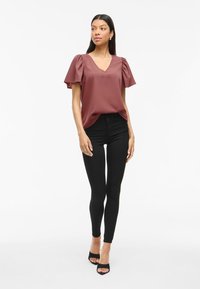 Blusa bordeaux con scollo a V, maniche a sbuffo, tessuto morbido; abbinata a jeans skinny neri e sandali neri open-toe. Sfondo semplice.