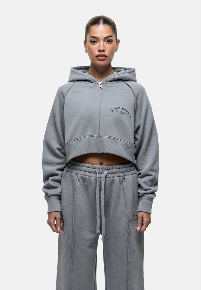 Felpa grigia a maniche lunghe con zip e ricamo frontale, abbinata a pantaloni da tuta grigi coordinati con vita regolabile tramite cordino e cuciture laterali.