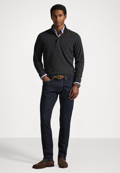 Σκούρος γκρίζος πουλόβερ με μισό φερμουάρ στον γιακά, συνδυασμένο με σκούρα μπλε τζιν και καφέ σουέντ loafers. Ένα ανοιχτό μπλε πουκάμισο φαίνεται από τον γιακά.