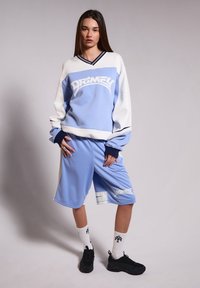 Suéter azul claro con acentos blancos y logotipo, combinado con pantalones cortos azul claro a juego. Zapatillas negras y calcetines blancos con texto negro.