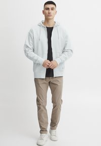 Lyseblå hettejakke med glidelås og snorhette, frontlommer, sammen med svart t-skjorte, beige bukser, og hvite joggesko.