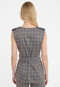 Mouwloze grijze jumpsuit met een patroon van witte rasterlijnen, ceintuurdetail op de taille en schouderaccenten; gemaakt van een gestructureerde stof.