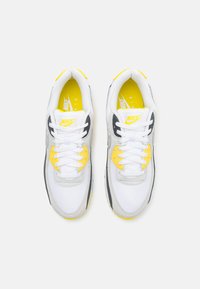 Zapatillas Nike en blanco y gris con detalles en amarillo, que cuentan con una parte superior texturizada, cordones redondos y una suela con amortiguación de aire visible.