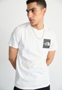 Balta medvilninė marškinėliai su juodu kvadratiniu logotipu, kuriame yra tekstas "THE NORTH FACE". Trumpomis rankovėmis, apvaliu kaklu, klasikinis siluetas.
