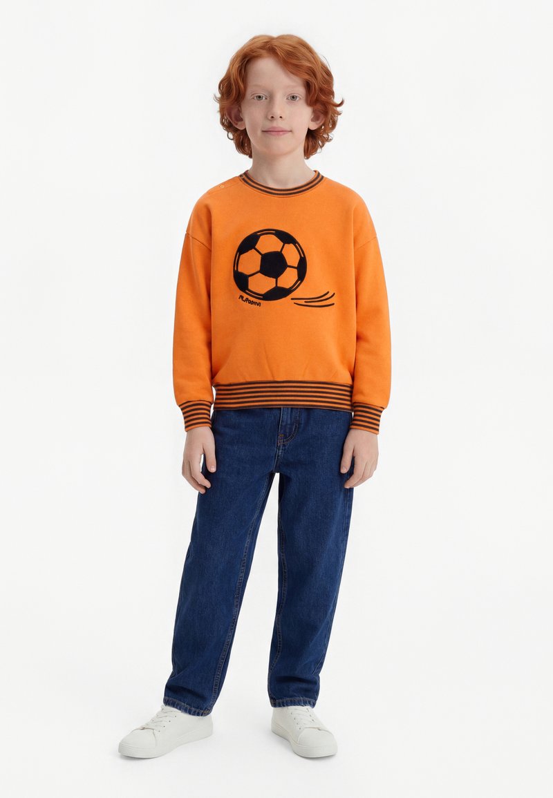 Niño con cabello rojo que lleva una sudadera naranja con un gráfico de balón de fútbol, jeans azules y zapatillas blancas, de pie contra un fondo blanco.