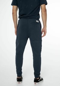 Marineblaue Jogginghose mit glatter Textur, rückseitiger Tasche, seitlichen Reißverschlüssen und schmalem Schnitt, kombiniert mit schwarzen Sneakers.