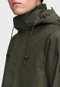 Vodoodolná, olivovo zelená bunda s vysokým golierom, nastaviteľnými šnúrkami a hladkou textúrou. Má minimalistický dizajn a žiadne viditeľné kovové prvky.