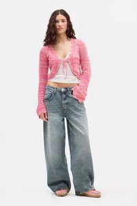 Cárdigan de punto rosa con un diseño corto y bordes en forma de concha, superpuesto a una blusa blanca. Combinado con jeans de pierna ancha azul claro y zapatos rosas.