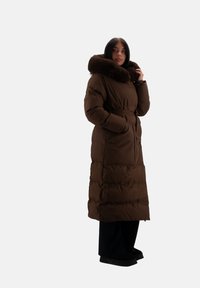 Cappotto imbottito marrone con un ampio colletto foderato in pelliccia, maniche lunghe, tasche laterali e vita con cinturino. Il design è ampio e arriva fino alla caviglia.