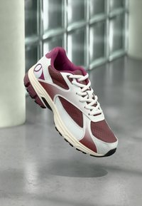 Chaussure de sport moderne avec des accents blancs, bordeaux et violets suspendue au-dessus d'un sol gris lisse près d'un mur en verre.