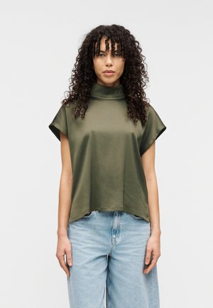 Olivgrünes Oberteil mit hohem Kragen, kurzen Ärmeln und lockerem Schnitt, gefertigt aus einem glatten Stoff. Kombiniert mit hellblauen Jeans mit weitem Bein.