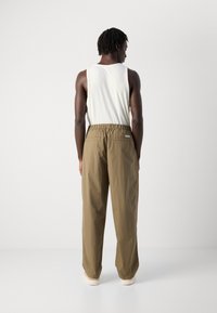 Man with dark skin and short dreadlocks stands facing a white wall, wearing a sleeveless white shirt, brown pants, and white shoes.