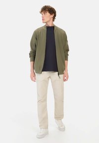 Olivenfarvet lynlåsjakke med ribbede manchetter, parret med en marineblå skjorte og lysebeige bukser, båret med hvide sneakers. Ensfarvet baggrund.