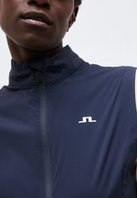 Κοντινό πλάνο ατόμου που φοράει σκούρο μπλε χωρίς μανίκια μπουφάν με φερμουάρ μπροστά και ένα μικρό λευκό λογότυπο στο στήθος.