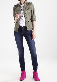 Olivengrøn jakke med skulderklapper, kombineret med en hvid grafisk t-shirt, mørkeblå skinny jeans og lyserøde ankelstøvler.