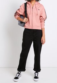 Pinkfarbener Kapuzenpullover mit Reißverschluss und schwarzen karierten Akzenten, kombiniert mit schwarzen hochgekrempelten Hosen und schwarz-weiß karierten Schuhen. Hält eine karierte Tasche.