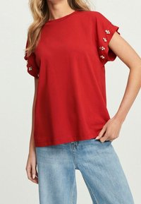 Vrouw in een rode shirt met korte mouwen, versierd met kleine, met edelstenen bezette bloemdetails op de mouwen, gecombineerd met lichtblauwe jeans.
