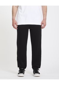 Pantalon de survêtement noir avec une coupe décontractée, des poignets élastiques et une texture lisse, associé à des baskets noires. Un T-shirt blanc recouvre la taille.