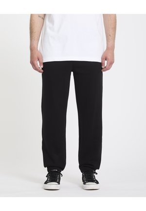 Pantalon de survêtement noir avec une coupe décontractée, des poignets élastiques et une texture lisse, associé à des baskets noires. Un T-shirt blanc recouvre la taille.
