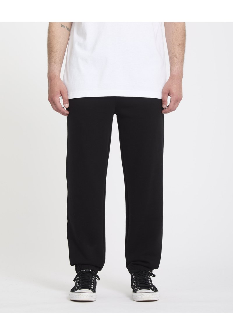 Pantalon de survêtement noir avec une coupe décontractée, des poignets élastiques et une texture lisse, associé à des baskets noires. Un T-shirt blanc recouvre la taille.