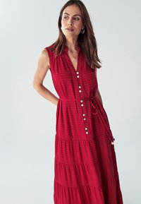 Červené šaty bez rukávov s golierom, zapínaním na gombíky a stiahnutým pásom. Vyrobené z textúrovanej tkaniny s tiered volánmi na sukni.